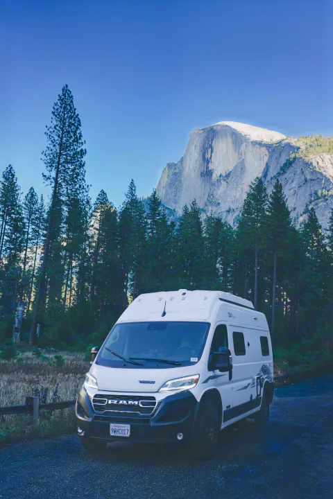 White rv rental camper van parked in Yosemite, with the towering Half Dome and tall pine trees under a clear blue sky.. Winnebago Winnebago 2026