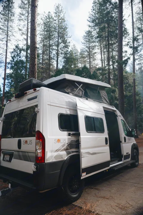 White Winnebago Solis camper van rv rental with pop-top deployed, nestled in a pine forest, ready for adventure.. Winnebago Winnebago 2026