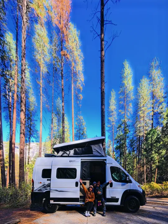Family enjoys their white Winnebago Solis rv rental, pop-top extended, amidst tall pine trees under a blue sky, experiencing nature.. Winnebago Winnebago 2026