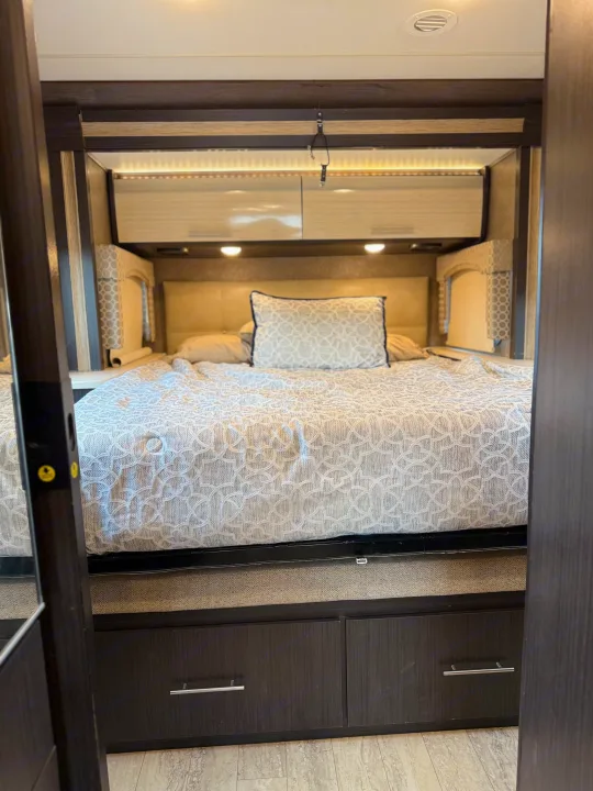 Comfortable RV rental bedroom featuring a bed with patterned bedding, overhead storage, ambient lighting, and built-in drawers. Ideal for travel.. Thor Axis 2018