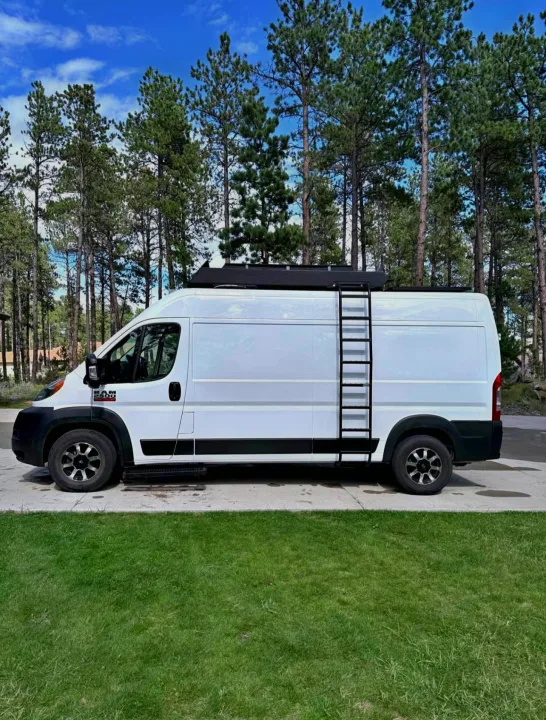 Explore with this white Ram ProMaster camper van rv rental, featuring a roof rack & ladder, parked by pine trees.. Ram Promaster 2500 2019