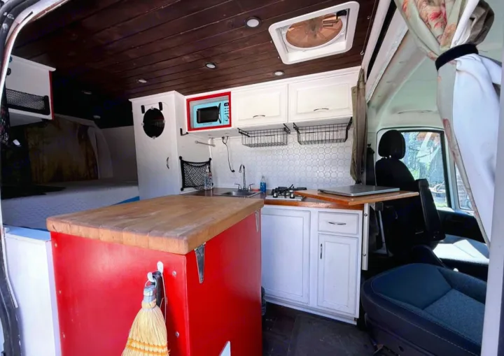 Custom rv rental interior: compact kitchen with light blue microwave, wooden ceiling, bed, and front seats.. Ram Promaster 2500 2019