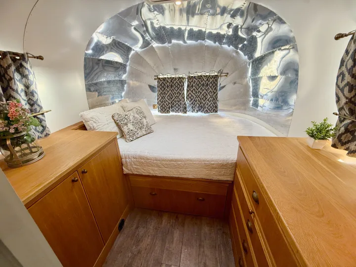 Cozy rv rental Airstream interior with shiny metal walls, a comfy bed, and warm wood cabinets on either side.. Airstream overlander 2018