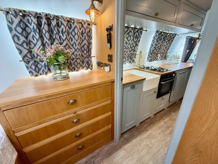 Cozy RV rental interior with a wooden dresser, floral decor, patterned curtains, and a compact kitchen featuring a farmhouse sink and oven.. Airstream overlander 2018