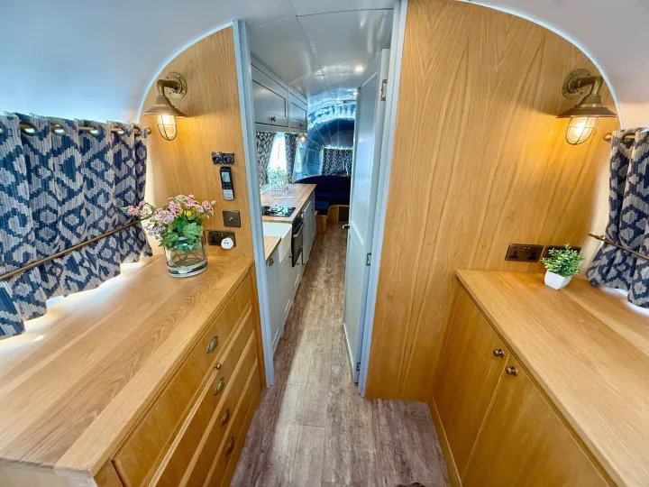 Step into this bright RV rental interior, featuring a sleek kitchen, warm wooden cabinetry, patterned curtains, and charming brass lights.. Airstream overlander 2018