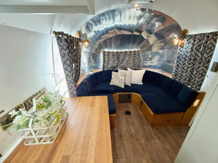 Inside a polished Airstream rv rental: U-shaped blue sofa, wooden table with flowers, patterned curtains, and bright lighting.. Airstream overlander 2018