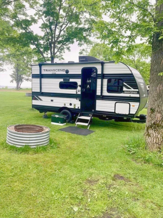 This RV rental, a Grand Design Transcend One, is set up at a grassy campsite with a fire pit, surrounded by trees.. Grand Design Transcend One 2025