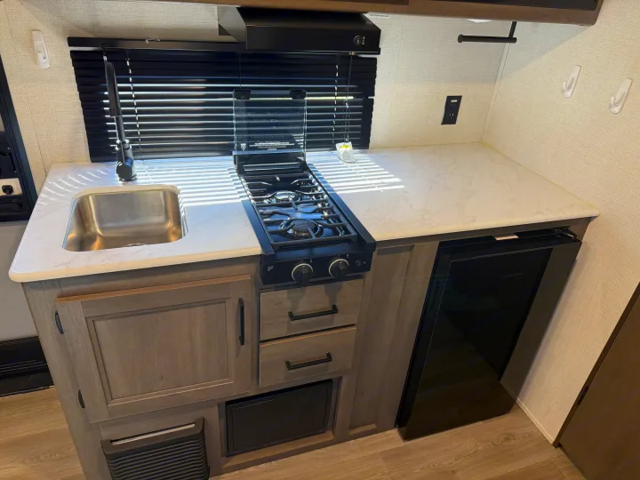 This RV rental kitchen features a sink, two-burner stove, mini-fridge, and light-colored countertops with cabinets below.. Grand Design Transcend One 2025