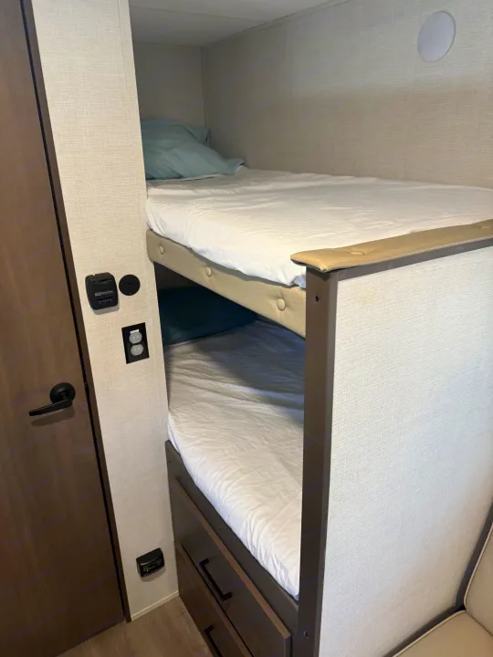 Inside an RV rental: view of stacked bunk beds with white sheets and pillows, integrated control panel and power outlets.. Grand Design Transcend One 2025