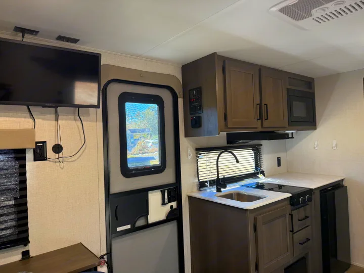 Step inside this RV rental, featuring a practical kitchen with sink, stove, microwave, cabinets, and a wall-mounted TV.. Grand Design Transcend One 2025