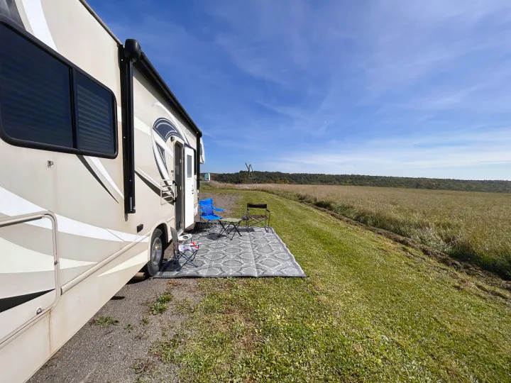 This tan RV rental, parked beside a vast field, offers a peaceful camp setup with chairs, rug, and scenic blue sky for relaxation.. Thor Motor Coach Chateau 2017