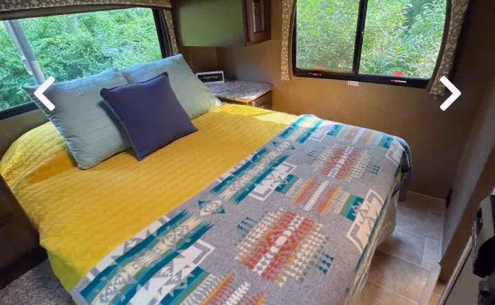 Cozy RV rental bedroom with a yellow comforter, patterned blanket, and pillows. Windows offer views of green foliage.. Thor Motor Coach Chateau 2017
