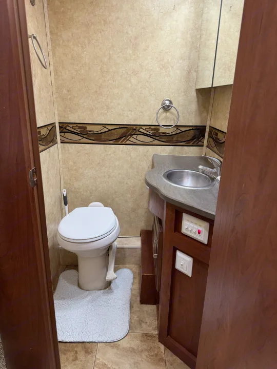 Compact RV rental bathroom with a white toilet, small sink, vanity, and light switches, on a tiled floor.. Jayco Melbourne 2012