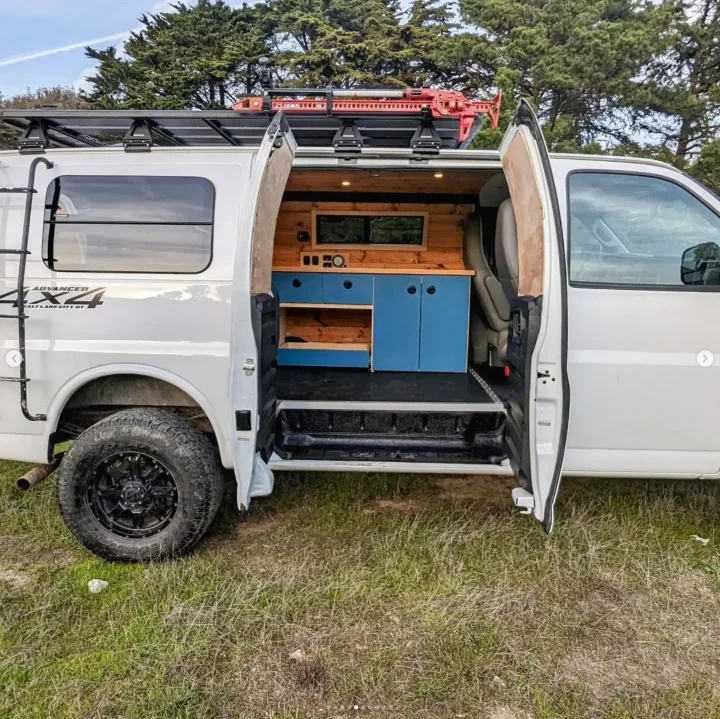 White off-road RV rental camper van. Open side reveals custom wood/blue interior, roof rack with jack & rugged tires. Adventure ready!. Chevrolet Express 2019