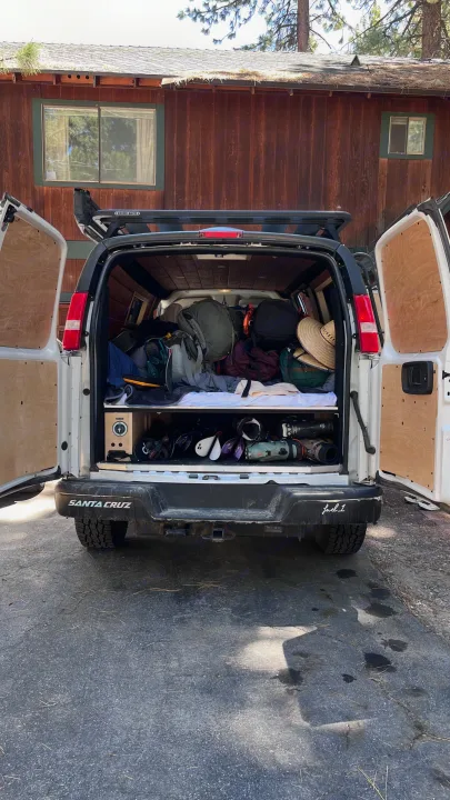 RV rental van, doors open, revealing a cozy, packed interior with outdoor gear, bags & a bed, parked near a cabin.. Chevrolet Express 2019