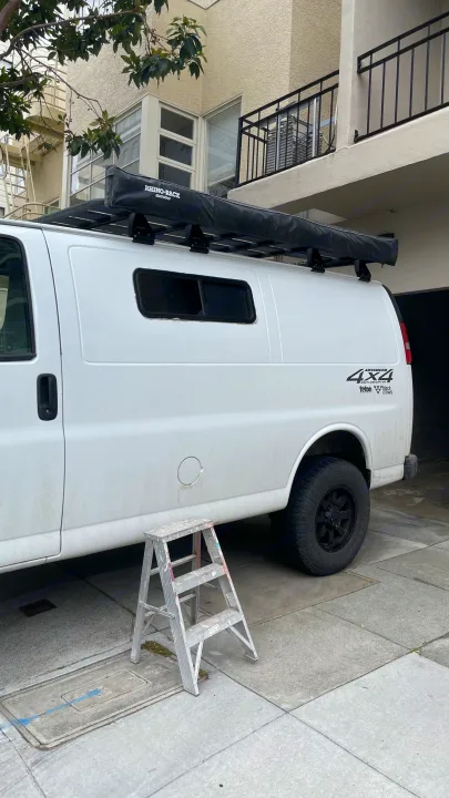 White 4x4 camper van (potential RV rental) with roof rack awning, parked. A metallic step ladder stands nearby.. Chevrolet Express 2019
