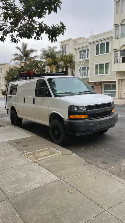 White 4x4 adventure van, potential RV rental, with roof rack & ladder, parked on a city street on an overcast day.. Chevrolet Express 2019