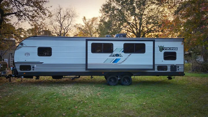 This large travel trailer, ideal for an rv rental, is set against autumn trees. Features include a slide-out & dual axles.. Forest River Viking 6K 2025
