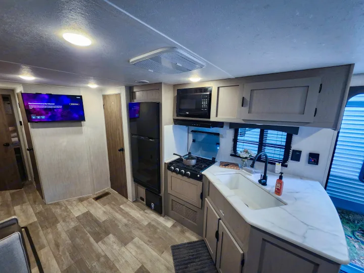RV rental kitchen and living area with stove, microwave, sink, and TV. Modern interior with wood-look flooring.. Forest River Viking 6K 2025
