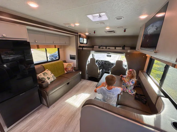 Two kids enjoy a spacious **RV rental** interior, featuring a lounge, kitchen, and TV. Perfect for family travel fun.. Forest River Coachmen Freelander 2026