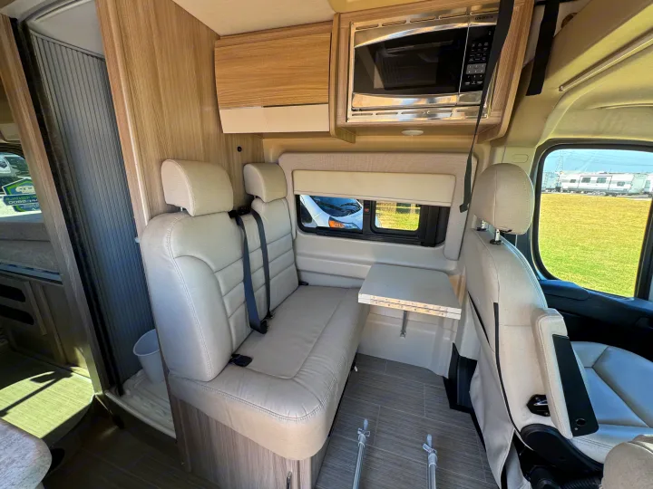 Inside an rv rental van: light-colored dinette seats, a wall-mounted microwave, and a folding table, ready for travel.. Thor Scope 2023