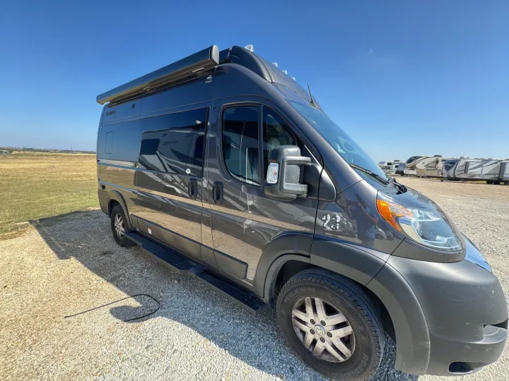A modern dark grey Class B camper van, ideal for rv rental, parked on gravel under a clear blue sky. Other RVs are in the background.. Thor Scope 2023