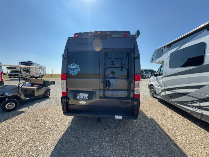 A black Thor Scope RV rental camper van is parked in a sunny gravel lot, surrounded by other RVs and a golf cart.. Thor Scope 2023