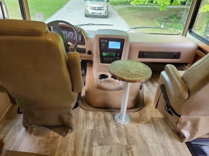 RV rental interior: cockpit with steering wheel, dashboard, driver and passenger seats, and a small table. An SUV is visible outside on a driveway.. Georgetown Georgetown Motorhome 2016