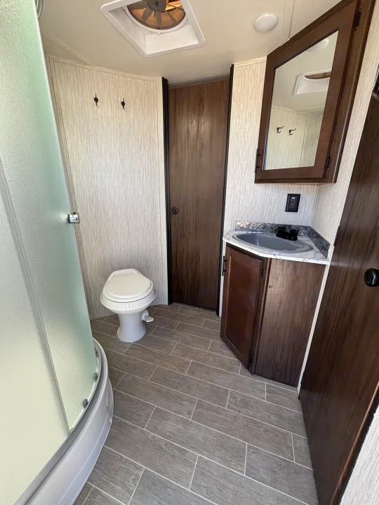 This RV rental bathroom features a compact toilet, frosted shower, sink vanity, and mirrored cabinet, all with wood-look flooring.. Forest River Cherokee Grey Wolf 2021