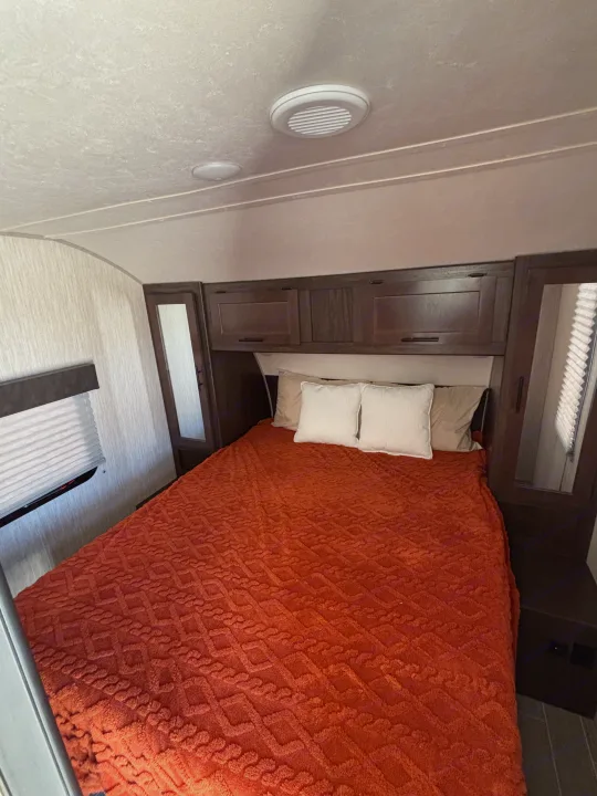 RV rental bedroom featuring a comfy bed with an orange blanket & pillows, overhead cabinets, and window. Ideal for travel.. Forest River Cherokee Grey Wolf 2021