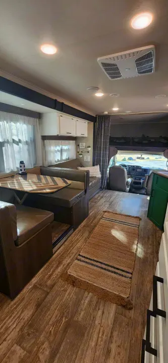 Modern rv rental interior with wood-look floors, a striped rug, dinette, and bright living space leading to the cab.. Jayco Redhawk 2017