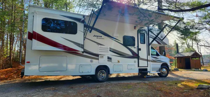 A white Jayco Redhawk rv rental, with its awning extended and side slide-out open, parked on a dirt path in a sunny forest.. Jayco Redhawk 2017