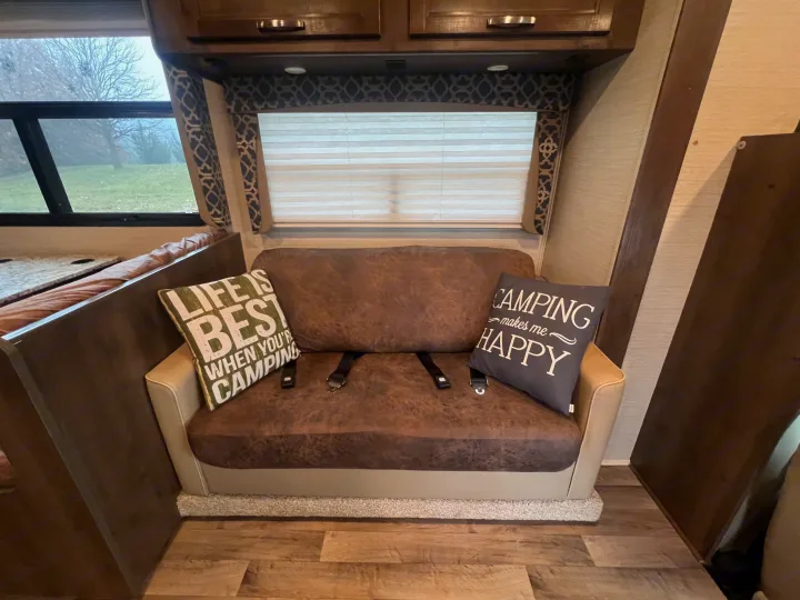 Cozy RV rental interior featuring a brown couch, camping pillows, window view of misty trees, and wood flooring.. Jayco Redhawk 2019