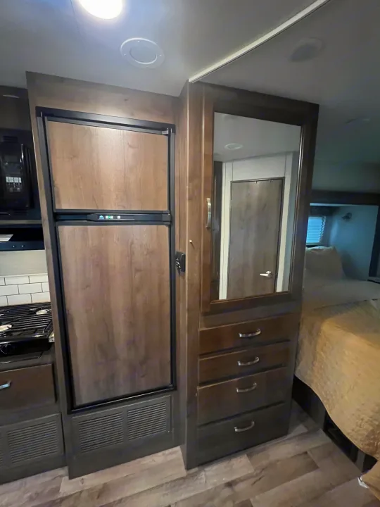 RV rental interior: Kitchenette with fridge, stove, microwave; adjacent mirrored storage dresser; and a glimpse of the bedroom.. Jayco Redhawk 2019