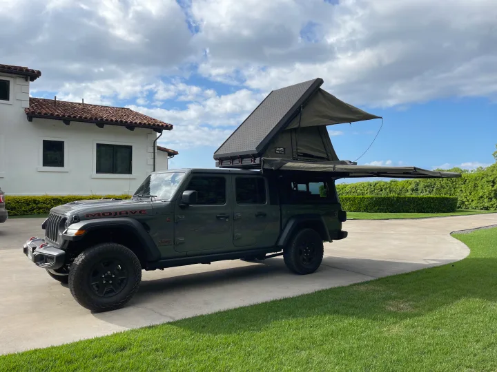 A dark green Jeep Gladiator, equipped with an open rooftop tent and side awning, parked on a driveway. Ideal for an RV rental adventure.. Jeep Gladiator 2022