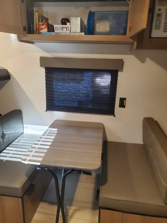 RV rental dinette setup: built-in benches, table, window with dark blinds, and open overhead storage filled with games & essentials.. Keystone Coleman 2026