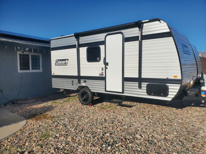 Bright white and grey Coleman travel trailer, perfect for an RV rental, parked on gravel next to a home on a sunny day.. Keystone Coleman 2026