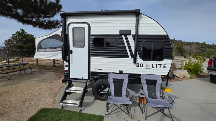Go Lite Skybreaker travel trailer rv rental set up at a scenic campsite. Features an extended pop-out, steps, and two camping chairs.. Wayfinder RV Go Lite 2025