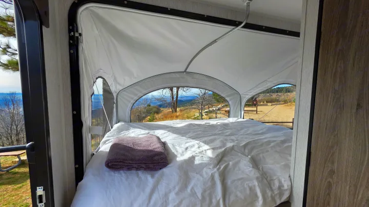 Cozy RV rental pop-up camper bed with a mountain view. White sheets, gray towel on bed. Perfect for scenic getaways.. Wayfinder RV Go Lite 2025