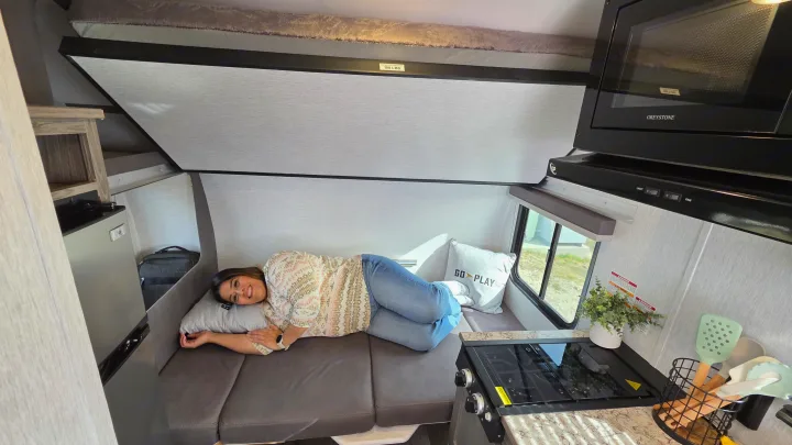 Woman relaxes inside a cozy RV rental, featuring a compact kitchen, microwave, and upper bunk bed.. Wayfinder RV Go Lite 2025