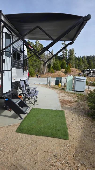 RV rental at a scenic campsite with an extended awning, chairs, and steps ready for relaxation amidst pine trees.. Wayfinder RV Go Lite 2025
