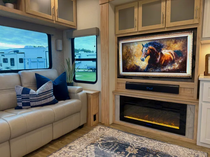 Inside an RV rental: a cozy living room with a cream couch, electric fireplace, TV displaying a horse, and outdoor views.. Alliance RV Avenue 2023