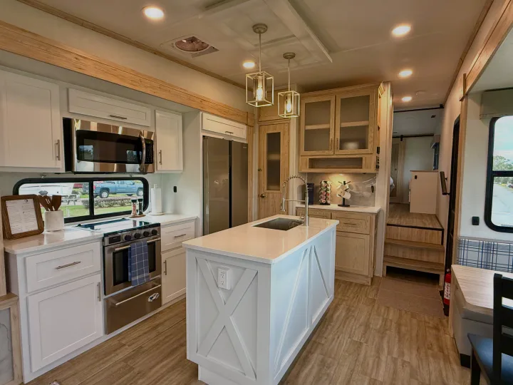 Modern RV rental kitchen featuring bright white cabinets, stainless steel appliances, a large island with a sink, and wood accents.. Alliance RV Avenue 2023