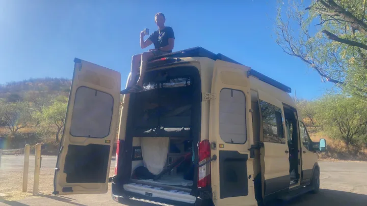 Man sips coffee on custom camper van roof, doors open, revealing interior. Your ultimate rv rental adventure awaits!. Ford Transit 2023