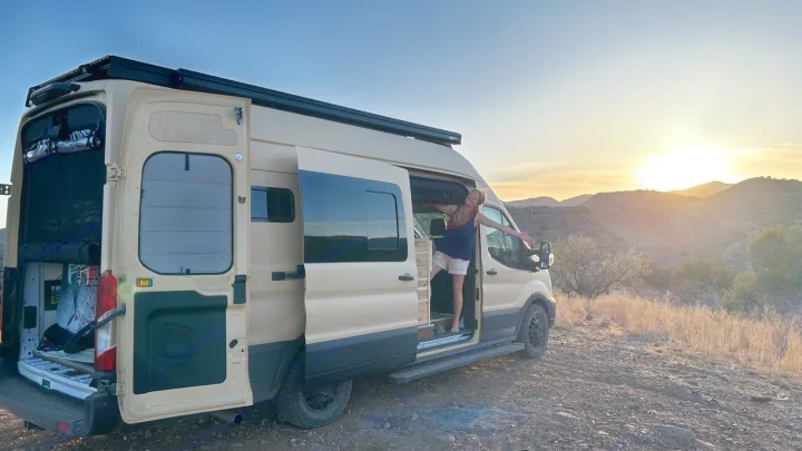 Smiling woman in her tan **RV rental** camper van, enjoying a beautiful sunset over scenic mountains. Perfect van life adventure!. Ford Transit 2023