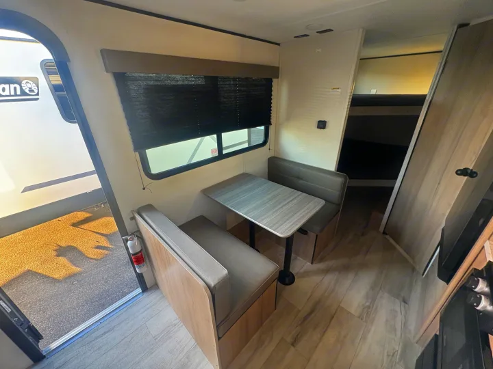 RV rental interior: light wood dinette with grey cushions, table, and bunk beds. Window with black blinds & fire extinguisher.. Keystone RV Coleman 2025