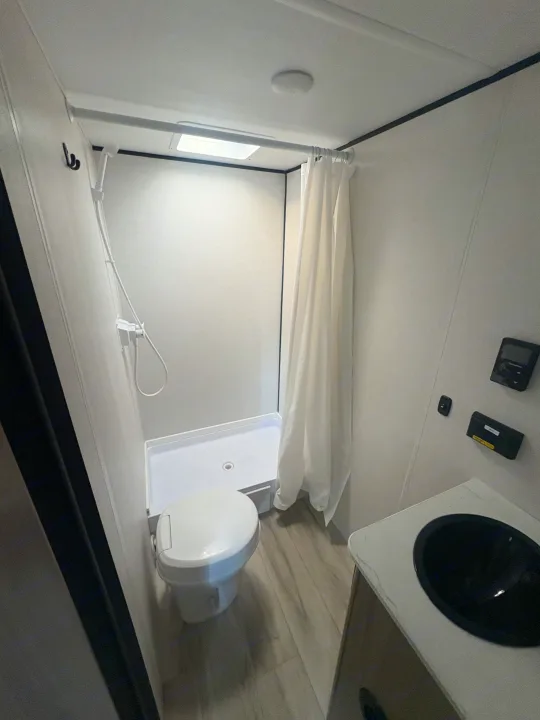 This compact RV rental bathroom features a toilet, shower with a white curtain, and a black sink for efficient travel living.. Keystone RV Coleman 2025