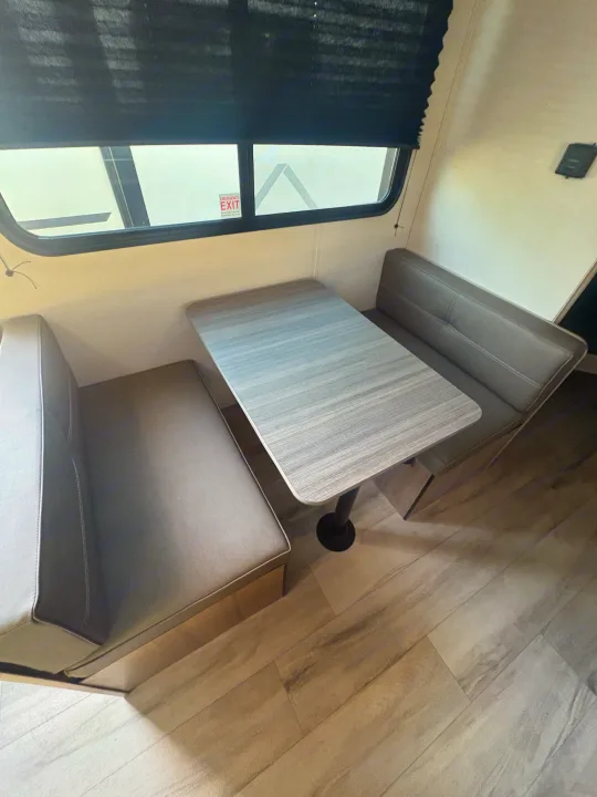 RV rental dinette featuring a wood-look table, grey bench seating, and window with an emergency exit.. Keystone RV Coleman 2025
