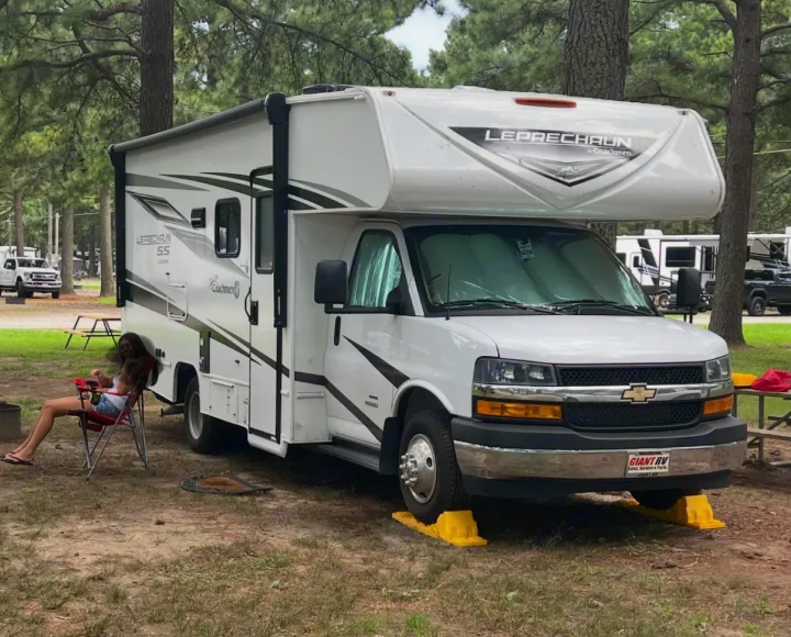 White Leprechaun rv rental motorhome parked on leveling blocks at a treed campsite, with a person relaxing in a chair.. Coachman Leprechaun 2025