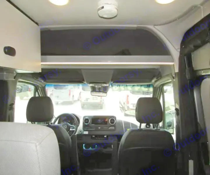 Interior of an RV rental van, looking toward the cockpit with overhead storage, front seats, and dashboard visible.. Winnebago Revel 2025